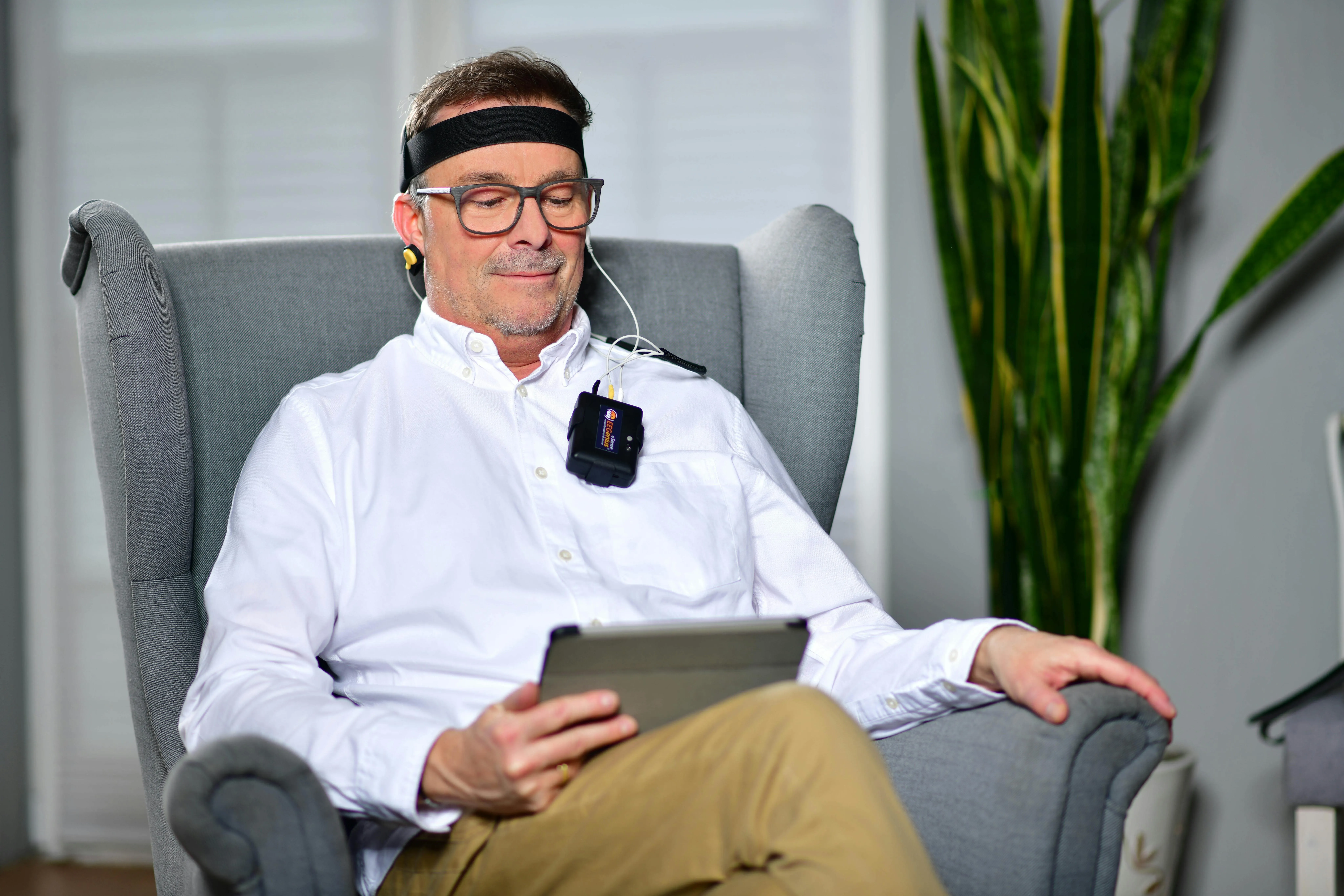 Man with EEG headband and biometric sensors reading a tablet in a chair